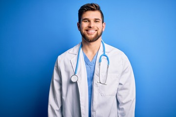 Young blond doctor man with beard and blue eyes wearing white coat and stethoscope with a happy and cool smile on face. Lucky person.