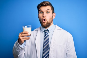 Young blond doctor man with beard and blue eyes wearing coat drinking glass of milk scared in shock with a surprise face, afraid and excited with fear expression
