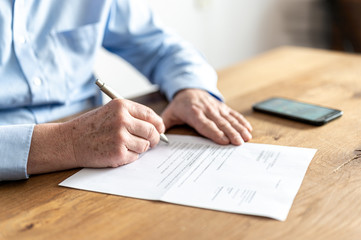 Hands of an older man with light blue shirt, sitting at a wooden table with a smartphone on it, while he is signing a letter.