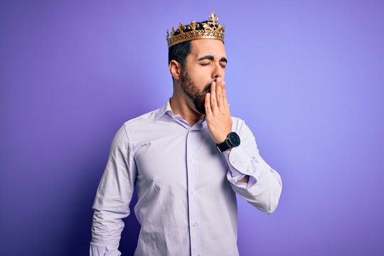 Young handsome man with beard wearing golden crown of king over purple background bored yawning tired covering mouth with hand. Restless and sleepiness.