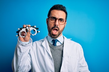 Young handsome optical man with beard holding optometry glasses over blue background In shock face, looking skeptical and sarcastic, surprised with open mouth