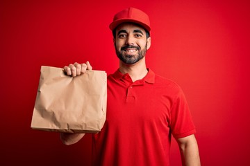 Young handsome delivery man with beard wearing cap holding takeaway paper bag with food with a happy face standing and smiling with a confident smile showing teeth