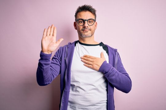 Young handsome man wearing purple sweatshirt and glasses standing over pink background Swearing with hand on chest and open palm, making a loyalty promise oath