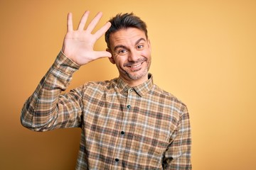 Young handsome man wearing casual shirt standing over isolated yellow background showing and pointing up with fingers number five while smiling confident and happy.