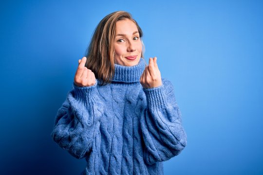 Young beautiful blonde woman wearing casual turtleneck sweater over blue background doing money gesture with hands, asking for salary payment, millionaire business