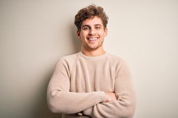 Young blond handsome man with curly hair wearing casual sweater over white background happy face smiling with crossed arms looking at the camera. Positive person.