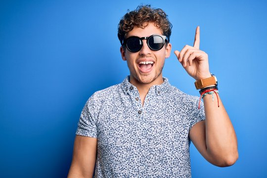 Young blond handsome man with curly hair on vacation wearing striped polo and sunglasses pointing finger up with successful idea. Exited and happy. Number one.