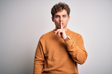 Young blond handsome man with curly hair wearing casual sweater over white background asking to be quiet with finger on lips. Silence and secret concept.