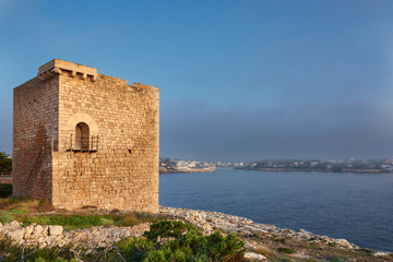 Fototapeta na wymiar Mediterranean landscape on a sunny, foggy morning. Medieval tower in the foreground and small fishing village in the background. Porto Petro, Mallorca. Balearic Islands. Spain.