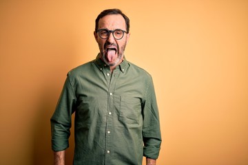 Fototapeta premium Middle age hoary man wearing casual green shirt and glasses over isolated yellow background sticking tongue out happy with funny expression. Emotion concept.