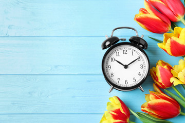 Black alarm clock with spring flowers and space for text on light blue wooden background, flat lay....