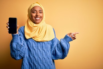 Young African American afro woman wearing muslim hijab showing smartphone very happy pointing with hand and finger to the side