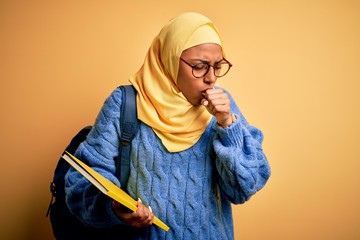 Young African American student woman wearing muslim hijab and backpack holding book feeling unwell and coughing as symptom for cold or bronchitis. Health care concept.
