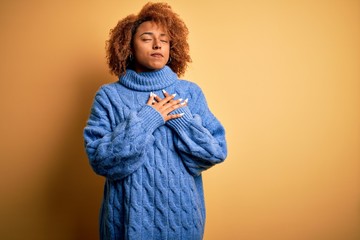 Young beautiful African American afro woman with curly hair wearing blue turtleneck sweater smiling with hands on chest with closed eyes and grateful gesture on face. Health concept.