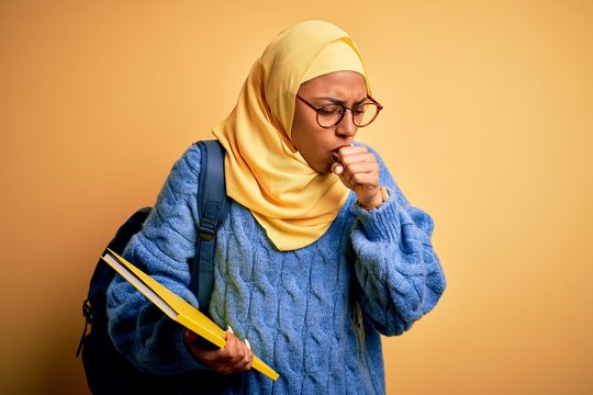 Young African American student woman wearing muslim hijab and backpack holding book feeling unwell and coughing as symptom for cold or bronchitis. Health care concept.