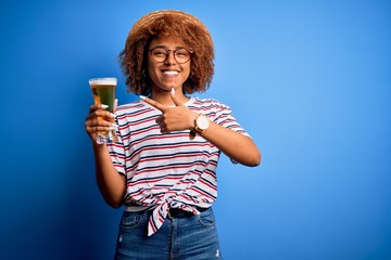 Young beautiful African American afro woman with curly hair on vacation drinking glass of beer very happy pointing with hand and finger