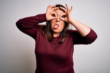 Beautiful brunette plus size woman wearing casual sweater over isolated white background doing ok gesture like binoculars sticking tongue out, eyes looking through fingers. Crazy expression.