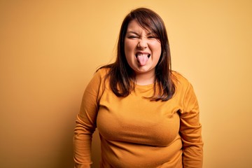 Obraz premium Beautiful brunette plus size woman wearing casual sweater over isolated yellow background sticking tongue out happy with funny expression. Emotion concept.