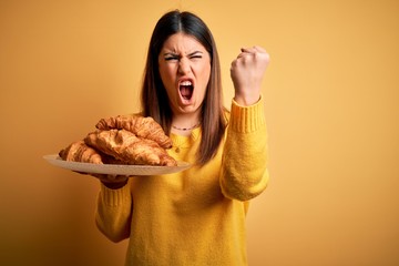 Young beautiful woman holding french croissant pastry over yellow background annoyed and frustrated shouting with anger, crazy and yelling with raised hand, anger concept