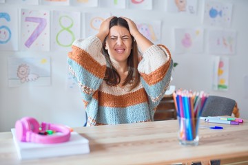 Young beautiful teacher woman wearing sweater and glasses sitting on desk at kindergarten suffering from headache desperate and stressed because pain and migraine. Hands on head.