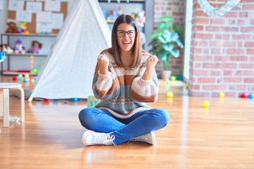 Young beautiful teacher woman wearing sweater and glasses sitting on the floor at kindergarten celebrating surprised and amazed for success with arms raised and open eyes. Winner concept.
