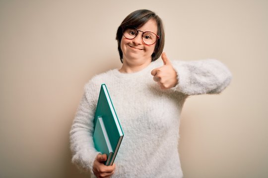 Young down syndrome student woman reading a book from library over isolated background happy with big smile doing ok sign, thumb up with fingers, excellent sign