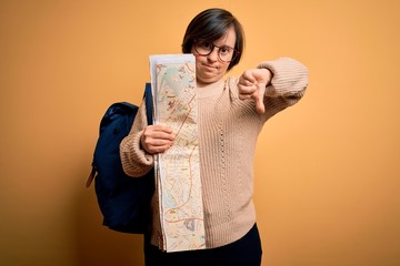 Young down syndrome tourist woman wearing bag and looking at destination city map with angry face,...