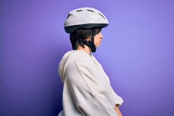 Young down syndrome cyclist woman wearing security bike helmet over purple background looking to side, relax profile pose with natural face with confident smile.