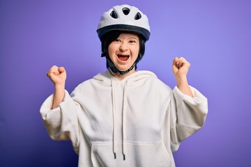 Young down syndrome cyclist woman wearing security bike helmet over purple background celebrating...