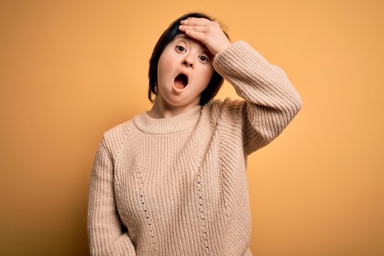 Young down syndrome woman wearing casual sweater over yellow background surprised with hand on head for mistake, remember error. Forgot, bad memory concept.