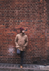 Young man with hat and warm clothes leaning against brick wall looking sideways in a London street