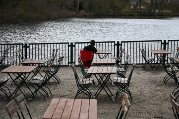 A young man sits alone on chair of an outside terrace in Berlin-Germany on a cold winter day while he looks at the lake.