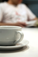 Close up shot of a white coffee cup and saucer, with an out of focus person in the background, reading a book