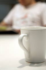 Close up shot of a white coffee mug, with an out of focus person in the background, reading a book