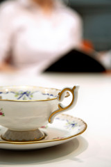 Obraz na płótnie Canvas Close up shot of an old fashioned china tea cup and saucer, with an out of focus person in the background, reading a book