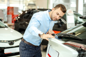 serious caucasian customer of car examining new car in dealership. guy in formal wear runs a finger over the surface of auto