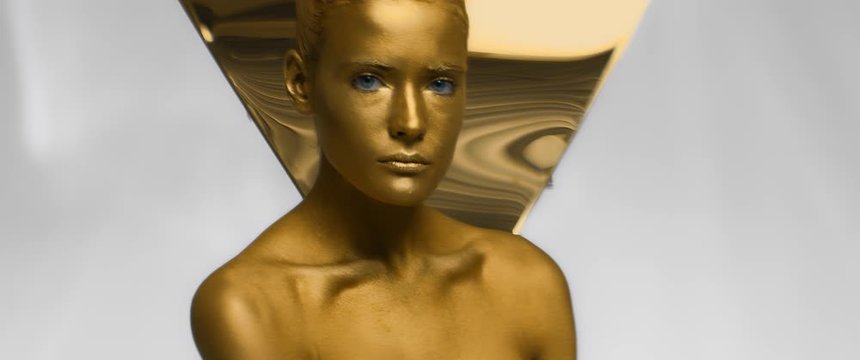 Gorgeous girl with blue eyes and golden covered skin poses looking at camera and touching neck and shoulder. Body art model woman with trendy shiny makeup in studio, decorated with metallic triangle