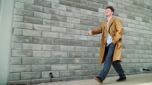 Stylish Beautiful Young Happy Caucasian Guy Goes along the City Wall and smiles. He is dressed in a Brown Long Coat. Close up. shooting on Steadicam - Powered by Adobe