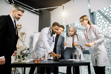 Multiethnic team of doctors, scientists, healthcare workers having a discussion, while using laptop, talking about the results of clinical trial or research