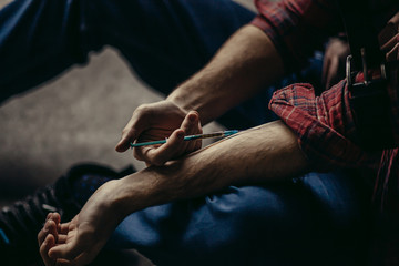 young caucasian man in red casual shirt injecting hard drugs into a veins with the use of syringe at first time, he embarked on a bad sinful path, being addicted by heroin - obrazy, fototapety, plakaty