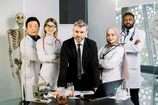Mature handsome man, medical professor smiling at the camera while standing at the table with group of his young multiracial students, doctors, lab workers. Laptop, microscope, test tubes and flasks