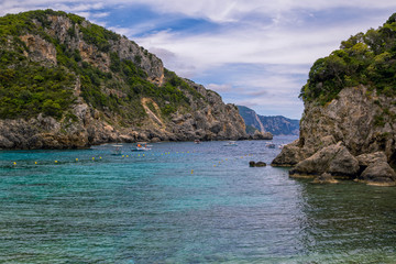 Fototapeta na wymiar Seascape – lagoon with turquoise water, mountain with cliffs, green trees, bushes, rocks in a blue water and fishers boats. Corfu Island, Greece. 