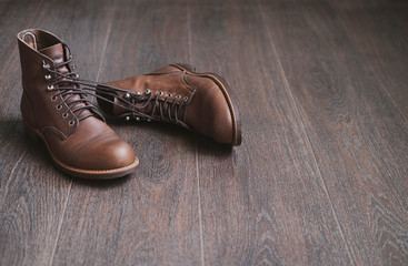 Two brown work leather men laced boots tied to each other on a wooden floor.
