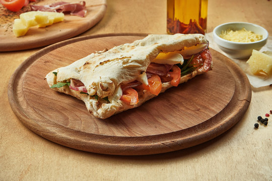 Close up view on tasty traditional italian sandwich panuozzo - oven-baked tortilla with tomatoes, melted mozzarella, prosciutto and arugula on a wooden board. Street food