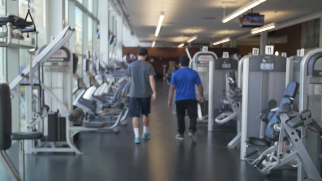 Two men Walk away in Gym surrounded by exercise equipment, trainer and client