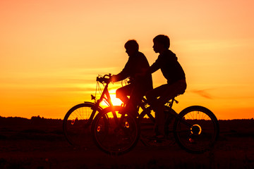 Fototapeta na wymiar Boy and senior woman riding bikes, silhouettes of riding persons at sunset in nature
