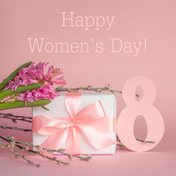 White gift box with pink ribbon, hyacinth flower, pussy-willow twigs and number 8 on pink background with text. Happy International Women's Day March 8 greeting card. Square image.
