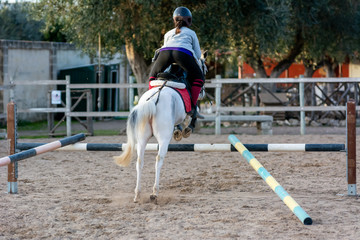 Back side of Girl riding a white horse jumping a guided obstacle during equestrian school training