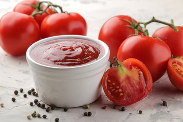 Red sauce or ketchup in a bowl and ingredients for cooking, spices, tomatoes and herbs on a light concrete background.