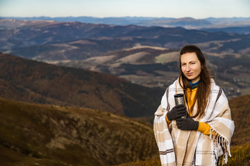 Naklejka premium Scenery autumn sunny day. Woman standing covered in plaid with termo cup in hand watching the mountains peak, sky with clouds.